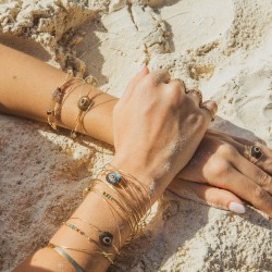 Photo artistique au soleil de l'accumulation des joncs fins Dulce et de la bague Triah sur une peau parsemée de grains de sable.