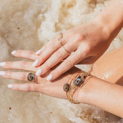 Focus sur les mains d'une femme posés sur le sable, portant la bague Triah et l'accumulation de joncs fins en or et grenat Enya.