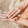 Focus sur les mains d'une femme posés sur le sable, portant la bague Triah et l'accumulation de joncs fins en or et grenat Enya.